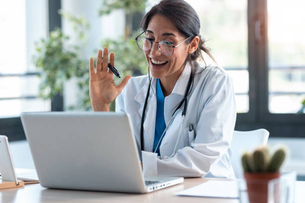 Shot of female doctor waving and talking with colleagues through a video call with a laptop in the consultation.